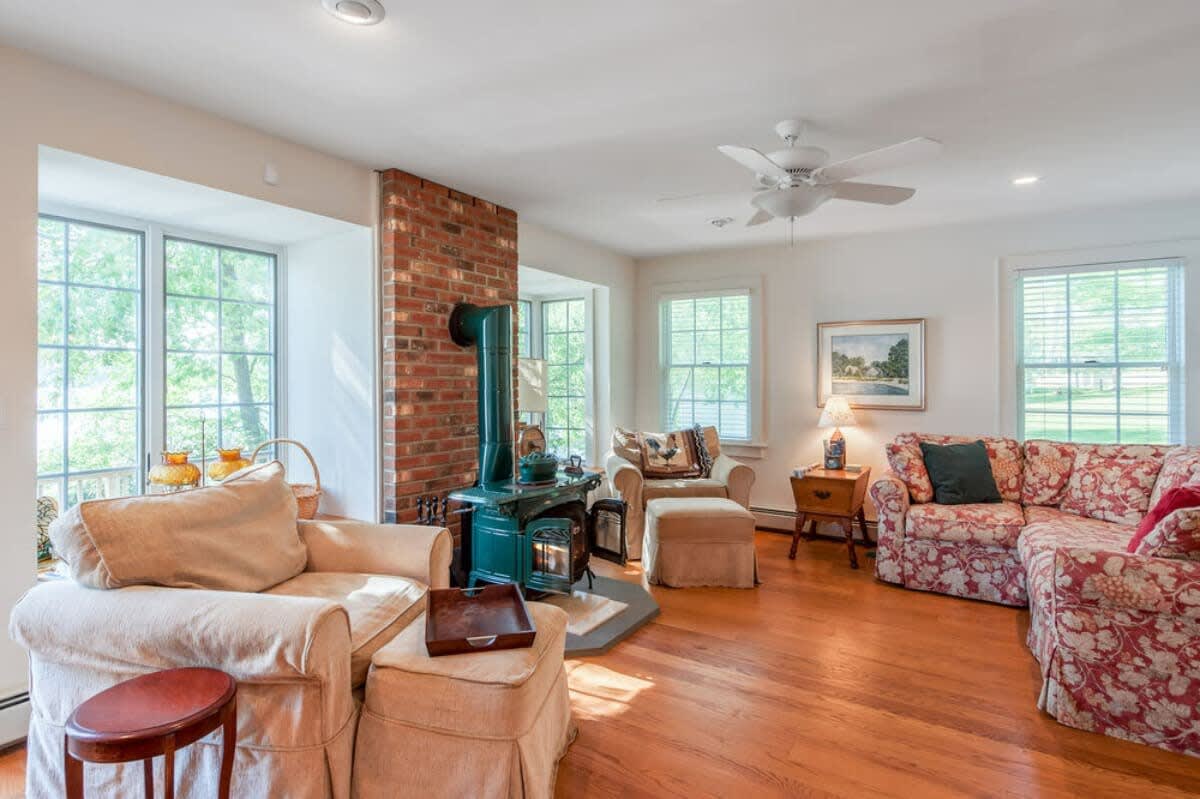 A spacious living room with polished hardwood floors and large windows, centered around a brick fireplace with a green wood-burning stove, and furnished with comfortable sofas and armchairs.