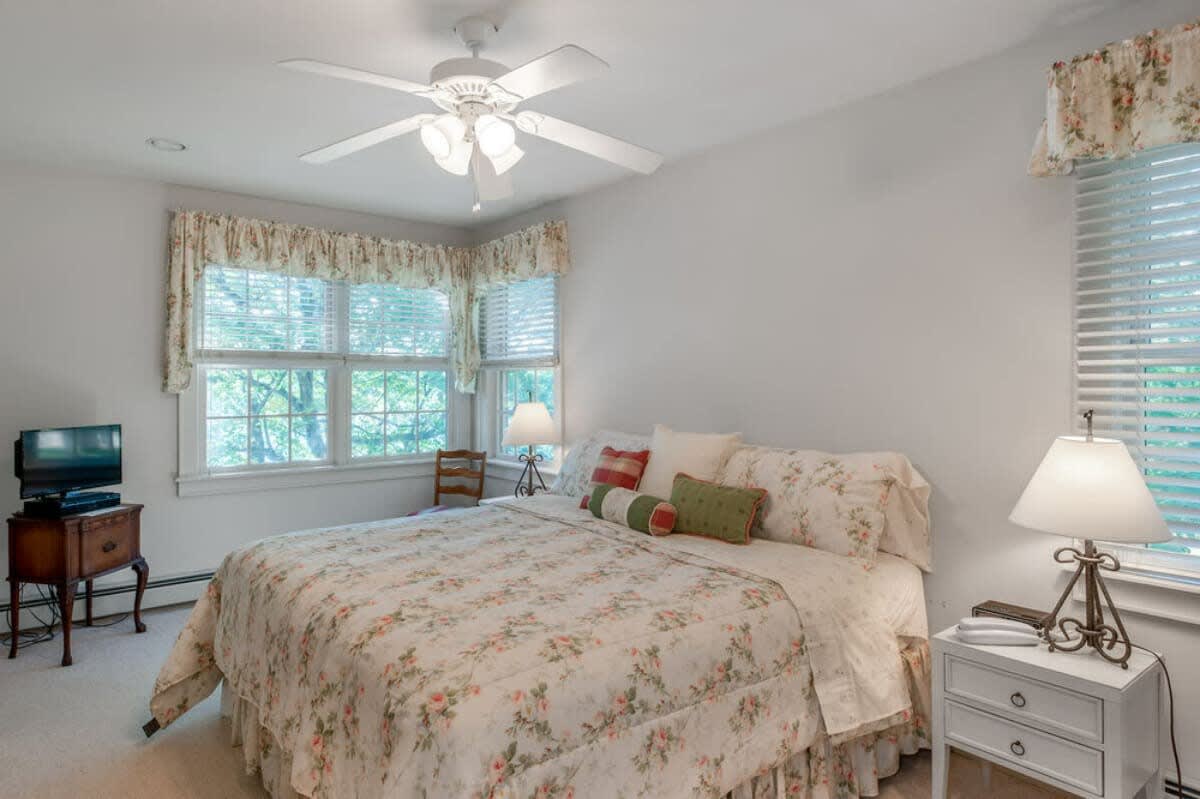 Comfortable bedroom with a floral-patterned queen bed, two large windows with blinds and valances, and a nightstand with a lamp, featuring a small TV on an antique table.