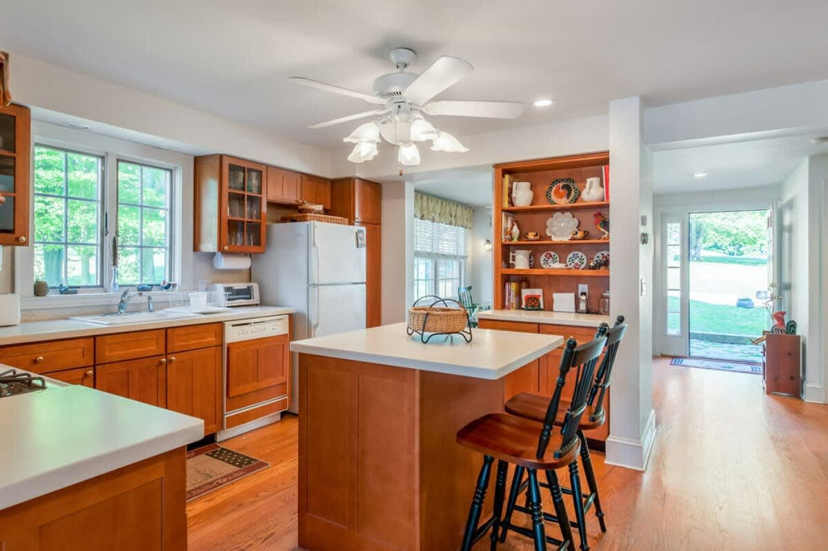 Spacious kitchen featuring light wood cabinets, white countertops, a central island with seating, and a built-in bookshelf, with hardwood floors extending towards an open doorway.