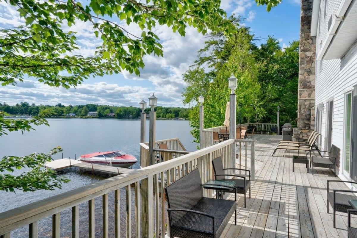 An expansive wooden deck with a railing, offering panoramic views of a serene lake with a boat docked at a private pier, surrounded by lush green trees.