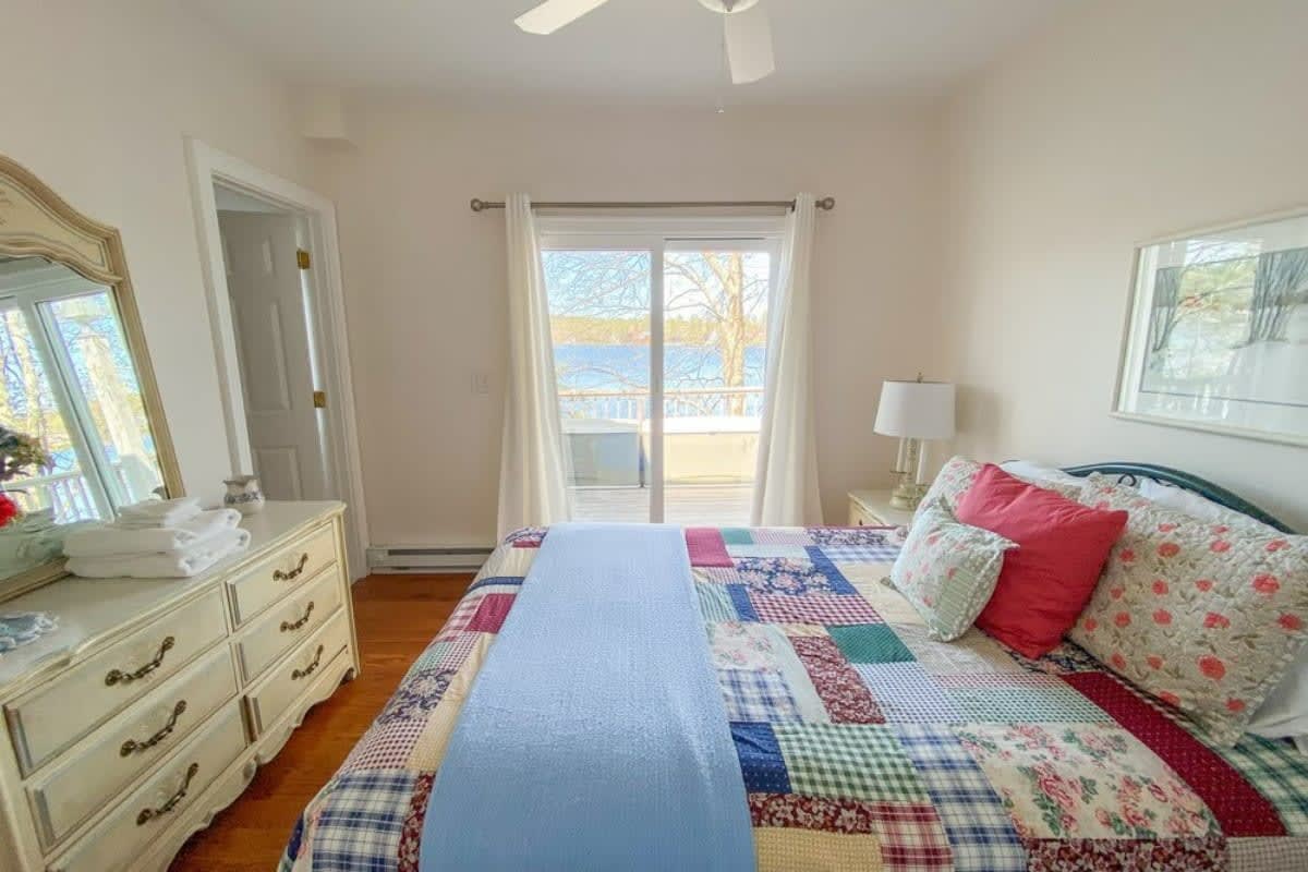 Bright bedroom featuring a large bed with a colorful patchwork quilt, a white dresser with a mirror, and sliding glass doors opening to a balcony with a scenic lake view.
