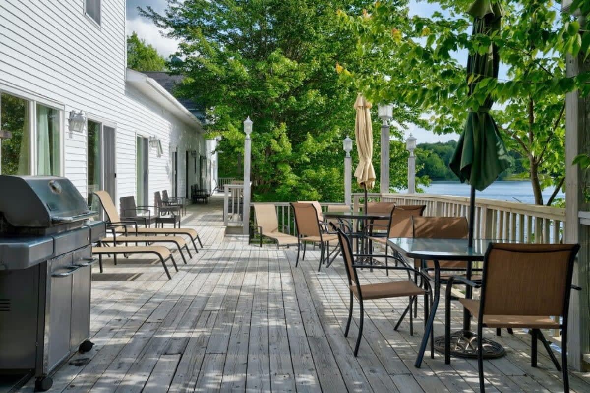 A spacious wooden deck furnished with multiple outdoor dining tables with umbrellas, lounge chairs, and a BBQ grill, all set against a backdrop of green trees and a lake.