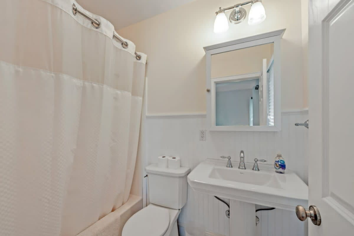 Clean bathroom with a white shower curtain over a bathtub, a white pedestal sink with a mirror above, and wainscoting on the walls, featuring a two-bulb vanity light.
