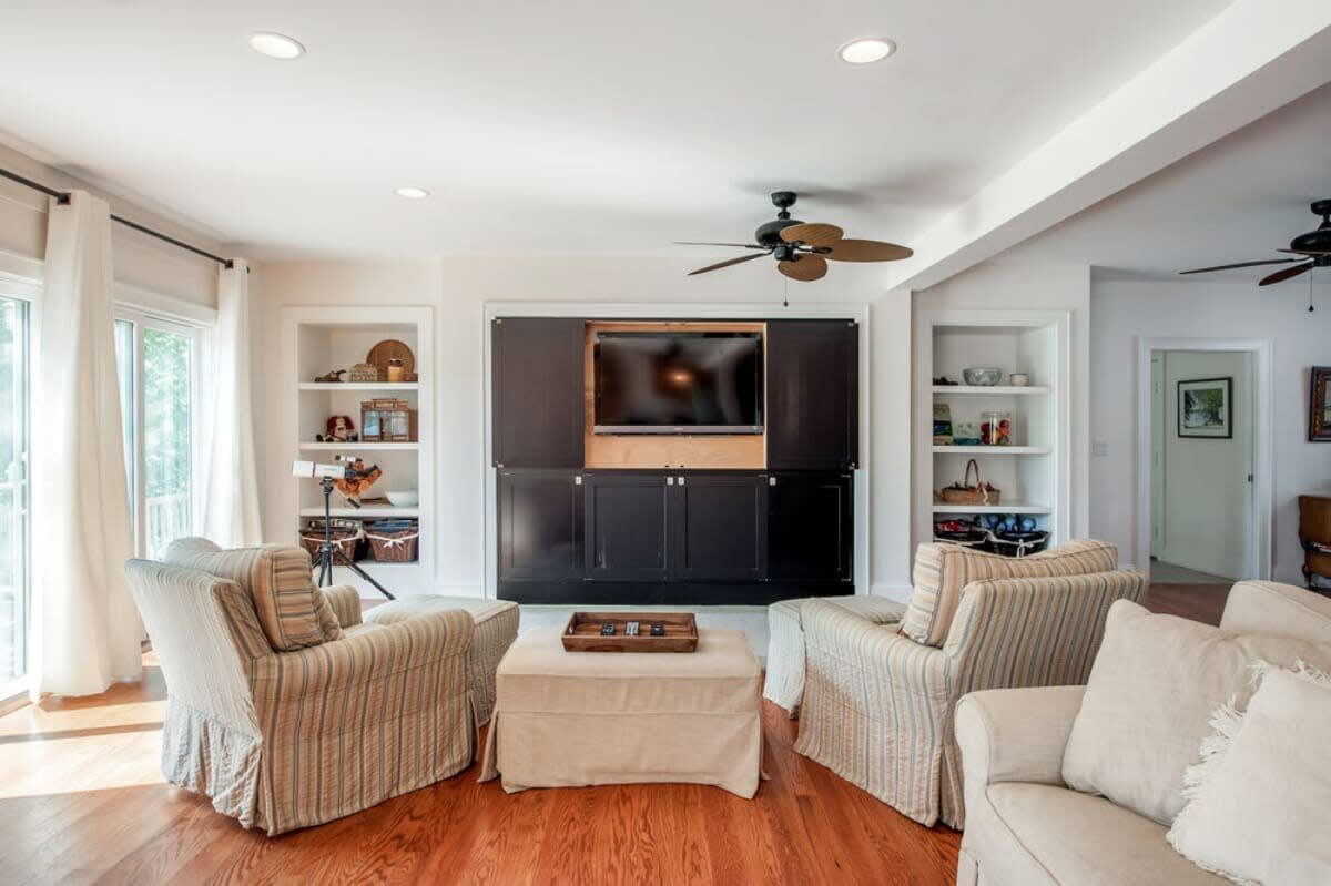 Living area with hardwood floors, two striped armchairs facing a dark entertainment cabinet with a TV, flanked by white built-in shelves and large windows.