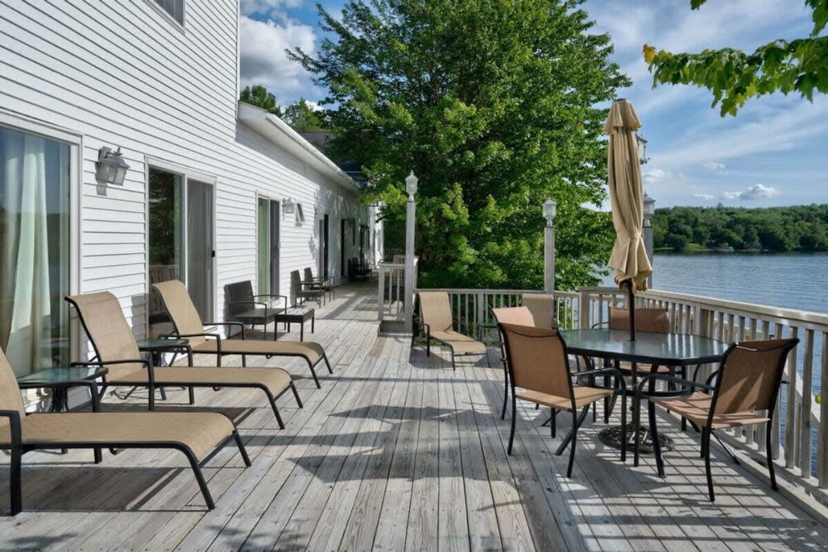 A spacious wooden deck extends along the back of the white house, furnished with multiple lounge chairs and a dining table with an umbrella, all offering expansive views of the serene lake. The deck is bordered by a white railing and lush greenery.
