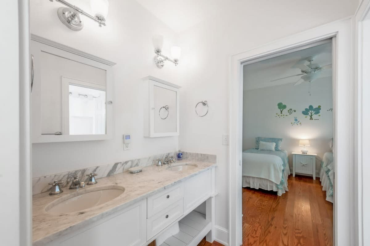 Modern bathroom featuring a double vanity with a marble countertop and two sinks, white cabinetry, and wall-mounted light fixtures. An open doorway reveals a glimpse into an adjacent bedroom.