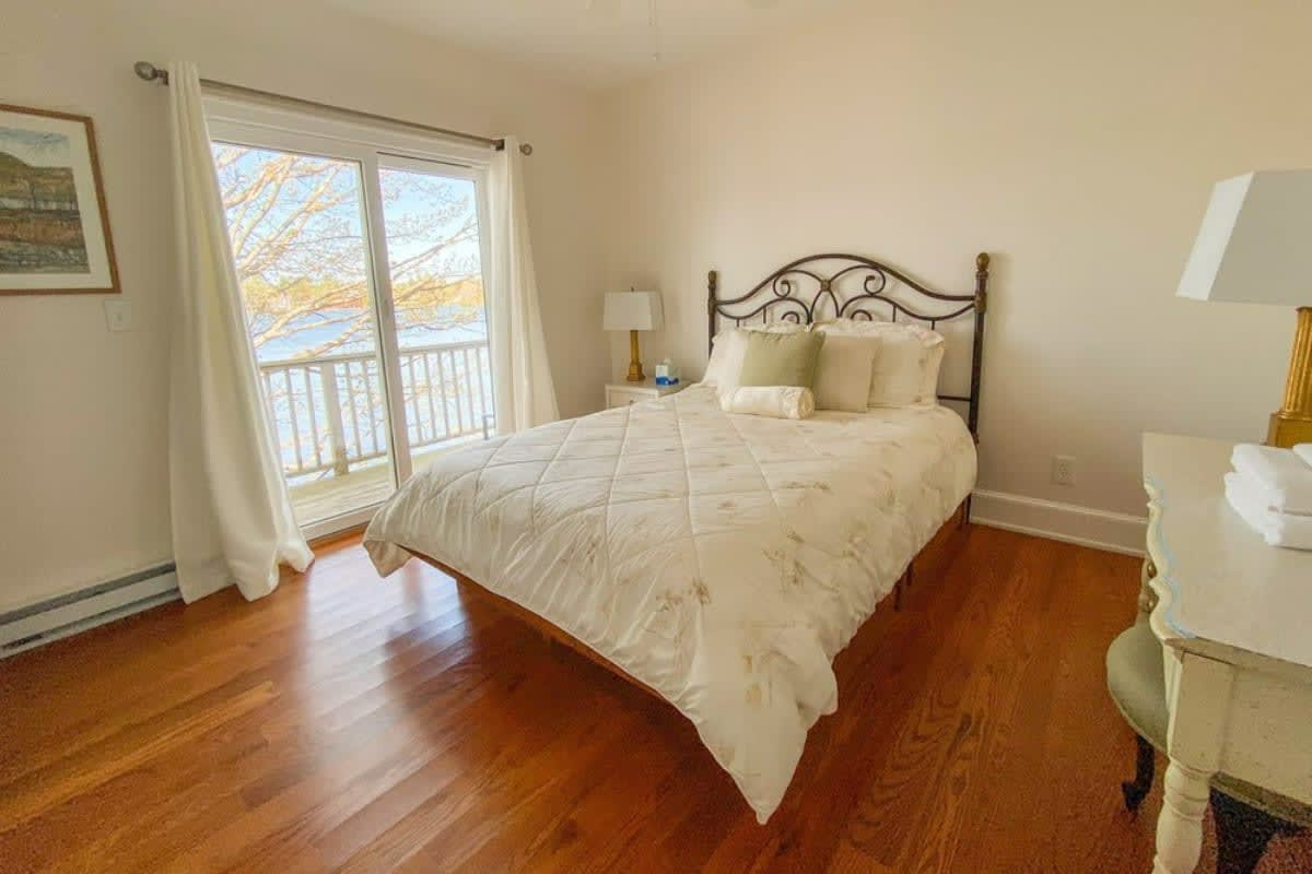 A cozy bedroom with polished hardwood floors, a wrought-iron queen-sized bed with white bedding, and a large sliding glass door leading to a balcony overlooking a lake.