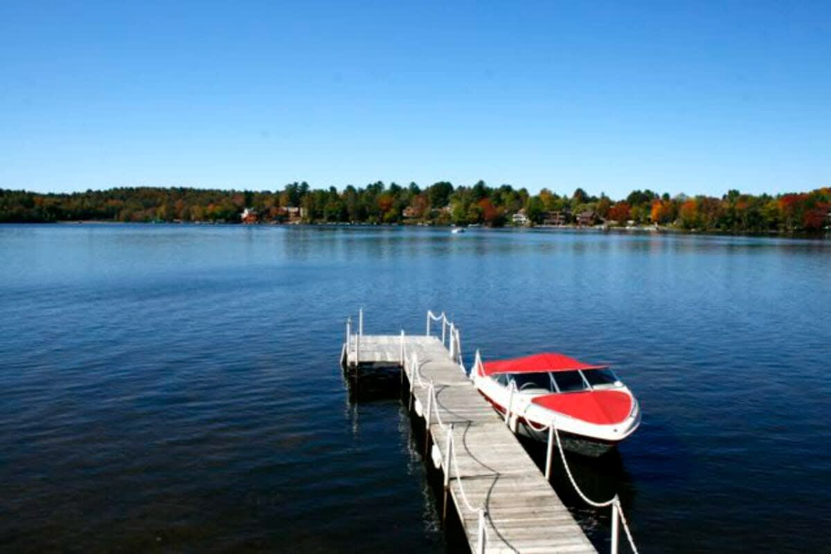 A picturesque view of a calm, blue lake with a long wooden dock extending into the water, where a red and white boat is moored, surrounded by distant tree-lined shores.