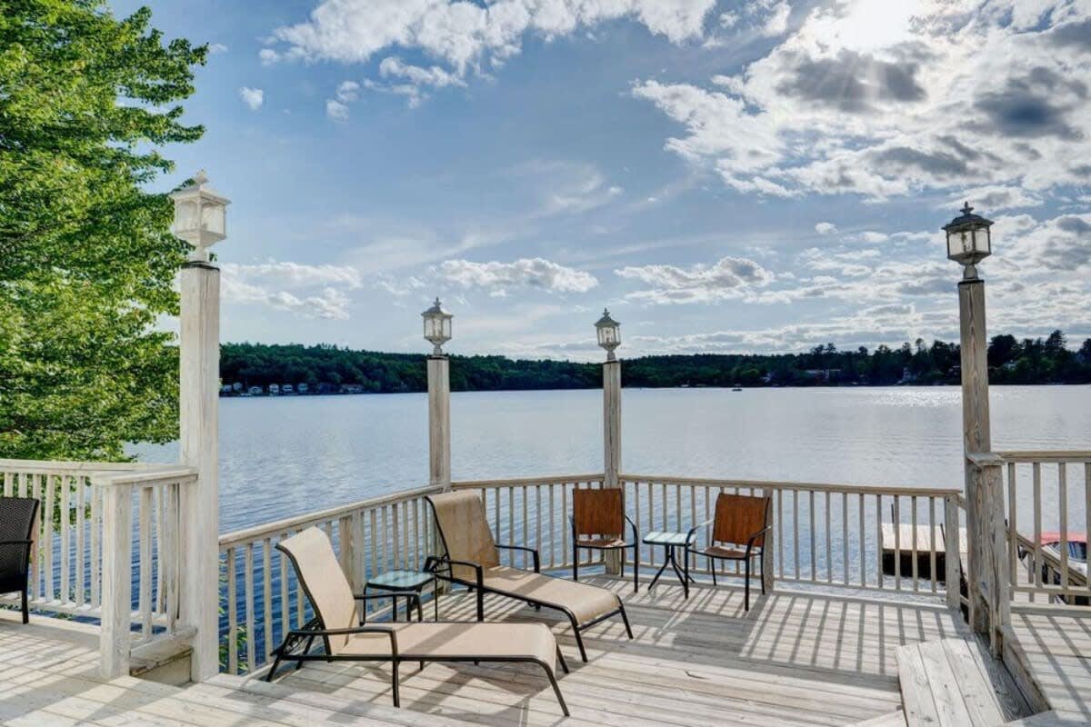 A wooden deck with white railings and decorative lanterns offers stunning panoramic views of a wide, calm lake surrounded by distant tree-lined shores. Lounge chairs and small tables invite relaxation in the sunny outdoor space.