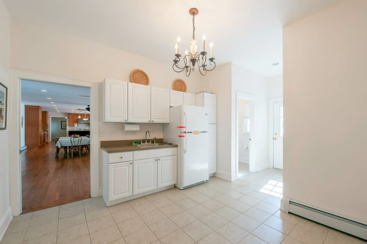 Compact secondary kitchen with white cabinetry, a sink, and a white refrigerator, set on tiled flooring, with an open doorway leading to another room and a chandelier light fixture.