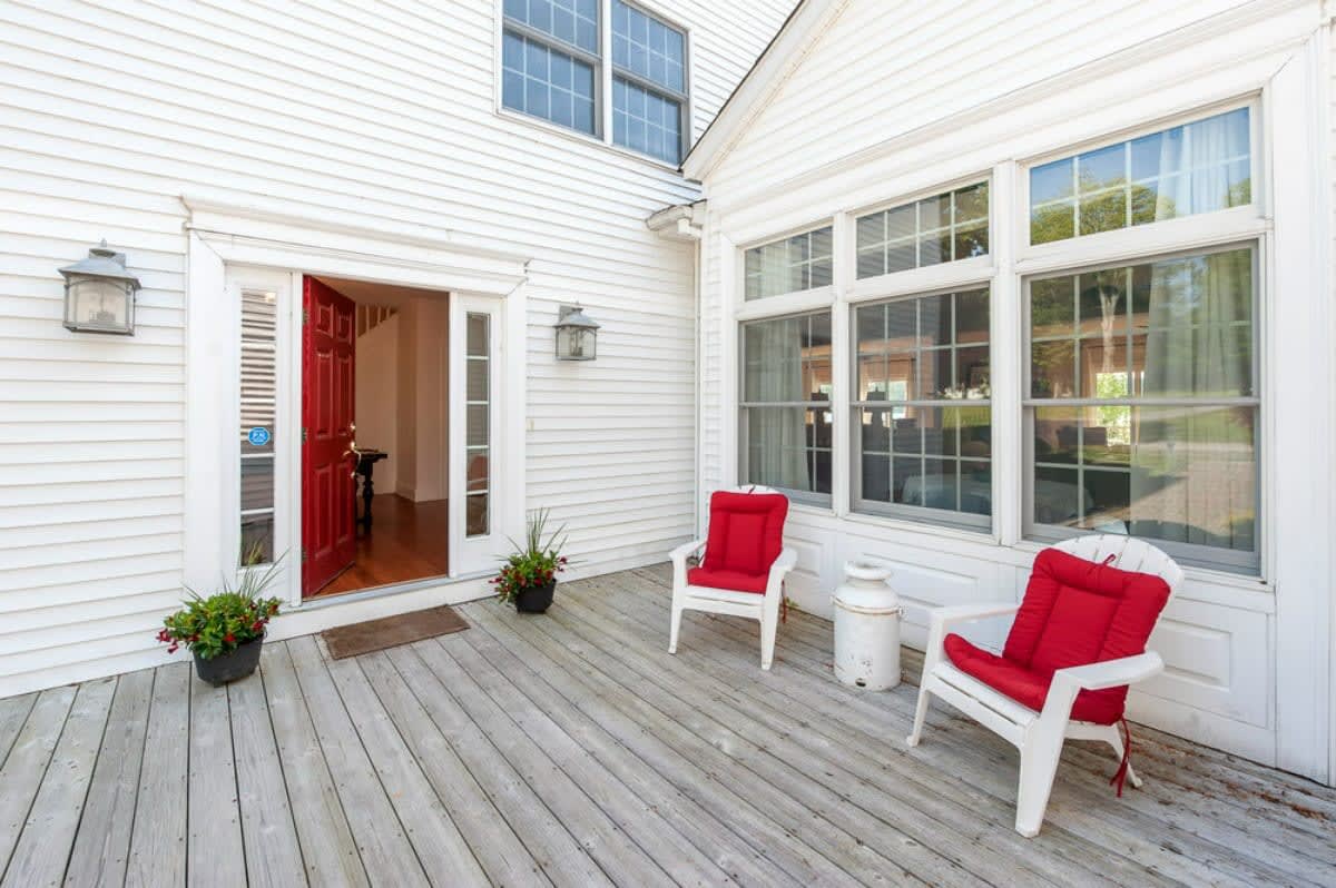 The front exterior of a white-sided house with a bright red front door, flanked by two white Adirondack chairs with red cushions on a wooden deck.