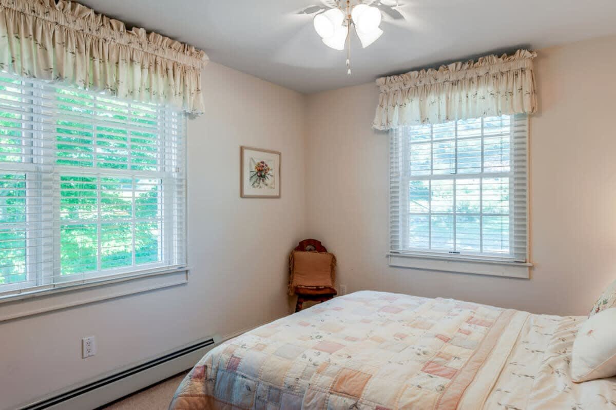 Cozy bedroom with a queen-sized bed covered in a patchwork quilt, two large windows with white blinds and valances, and light-colored walls, featuring a small decorative chair.