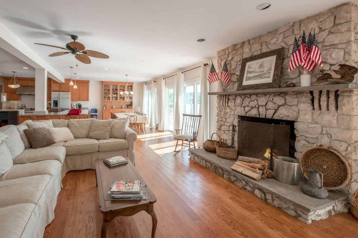 Cozy living room with a prominent stone fireplace, a large light-colored sectional sofa, and hardwood floors, decorated with American flags on the mantel.