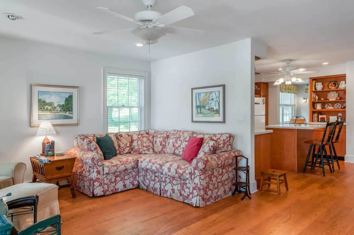 A bright living room with polished hardwood floors, featuring a floral patterned sectional sofa, a ceiling fan, and an open layout connecting to a kitchen with a breakfast bar.