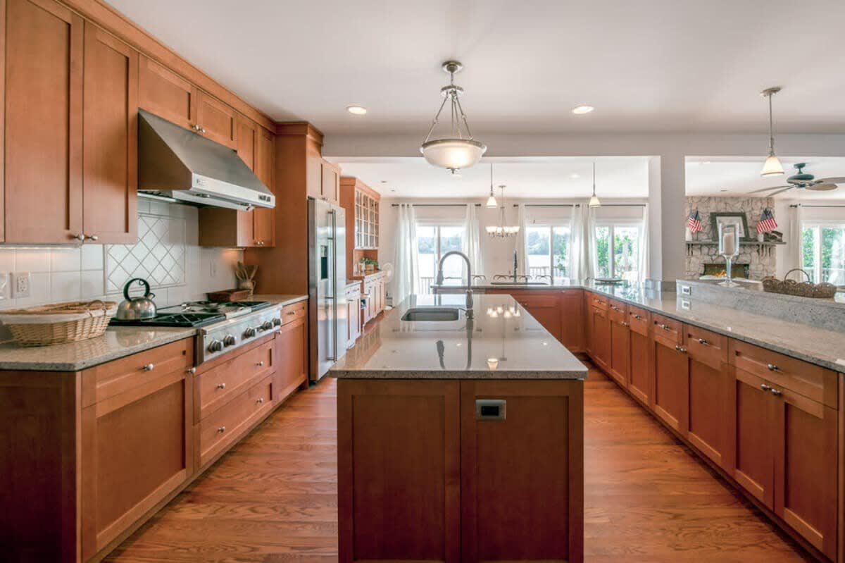 Expansive kitchen with light wood cabinets, granite countertops, a gas range with a hood, and a long central island featuring a sink and dishwasher, all on hardwood floors.