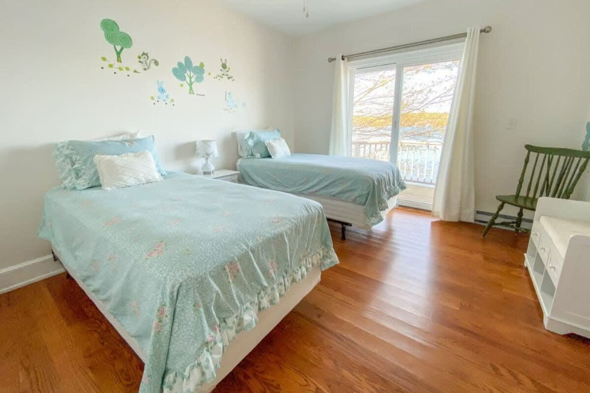 Cheerful children's bedroom with two twin beds featuring light blue bedding, set on polished hardwood floors. Playful tree and animal wall decals adorn the wall, and sliding glass doors open to a balcony with a lake view.
