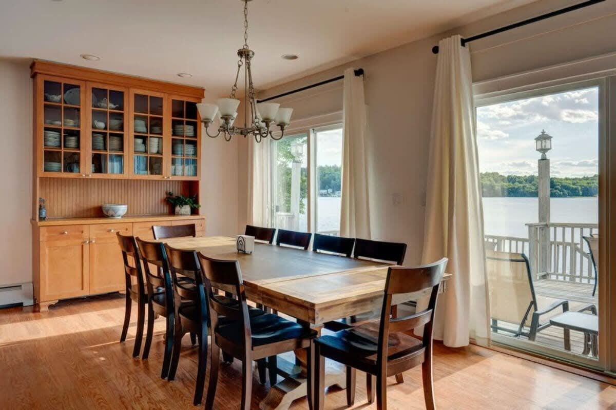 Bright dining room with a large wooden table, dark chairs, a built-in wooden hutch, and sliding glass doors leading to a deck with scenic lake views.