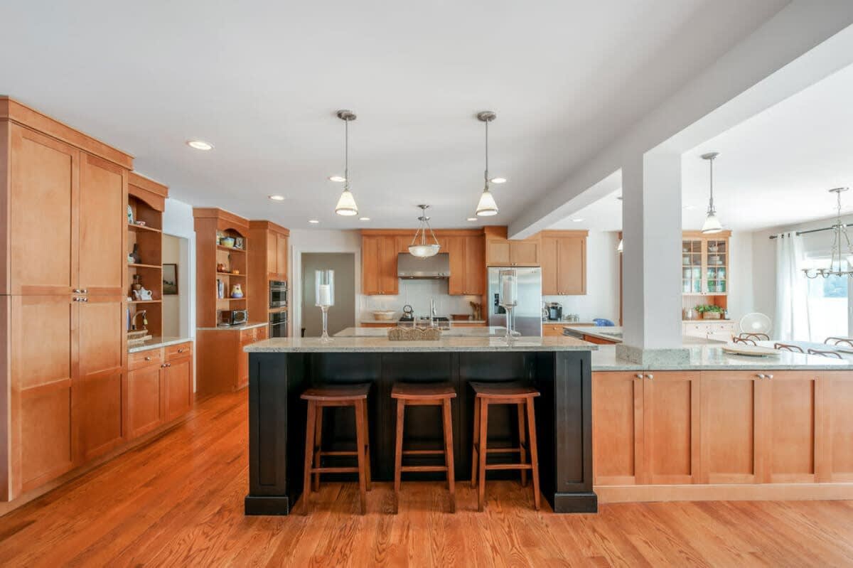 Spacious kitchen featuring light wood cabinetry, granite countertops, a large island with three bar stools, and stainless steel appliances, with hardwood floors extending into the open-concept space.