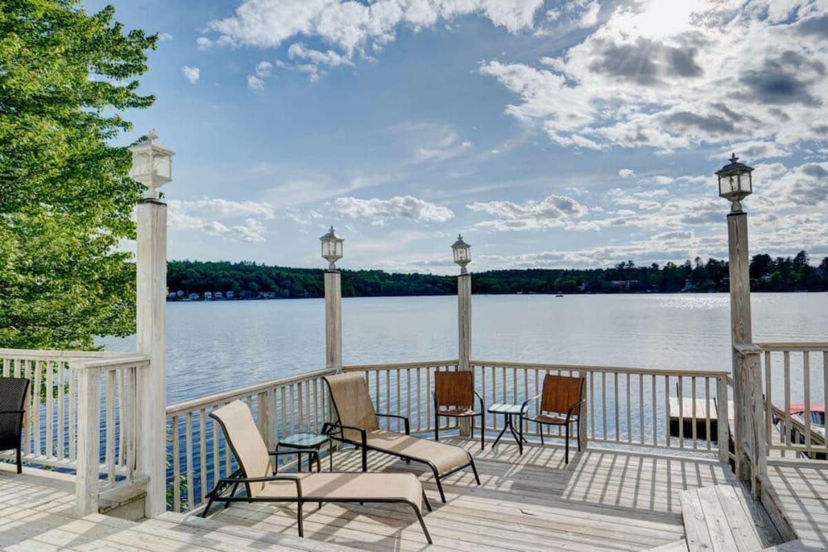 Expansive wooden deck furnished with lounge chairs and a small table, offering panoramic views of a wide lake and distant tree-lined shores under a partly cloudy sky. Decorative lanterns line the railing.