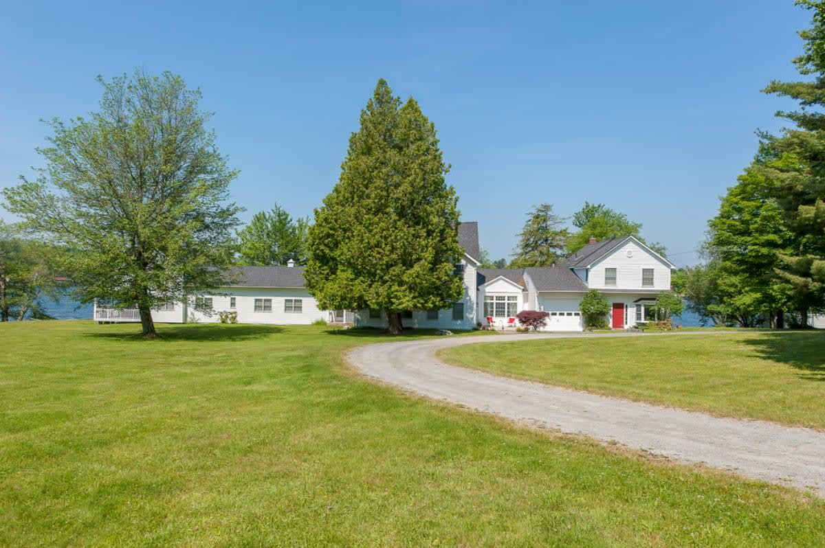 A long gravel driveway leading up to the white-sided house, surrounded by expansive green lawns and mature trees, with the lake visible in the background.