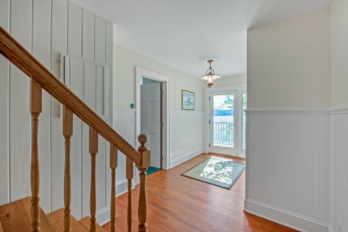 A bright hallway with polished hardwood floors and white beadboard wainscoting, featuring a wooden staircase railing on the left and a glass-paned door leading to an outdoor deck with lake views at the end.
