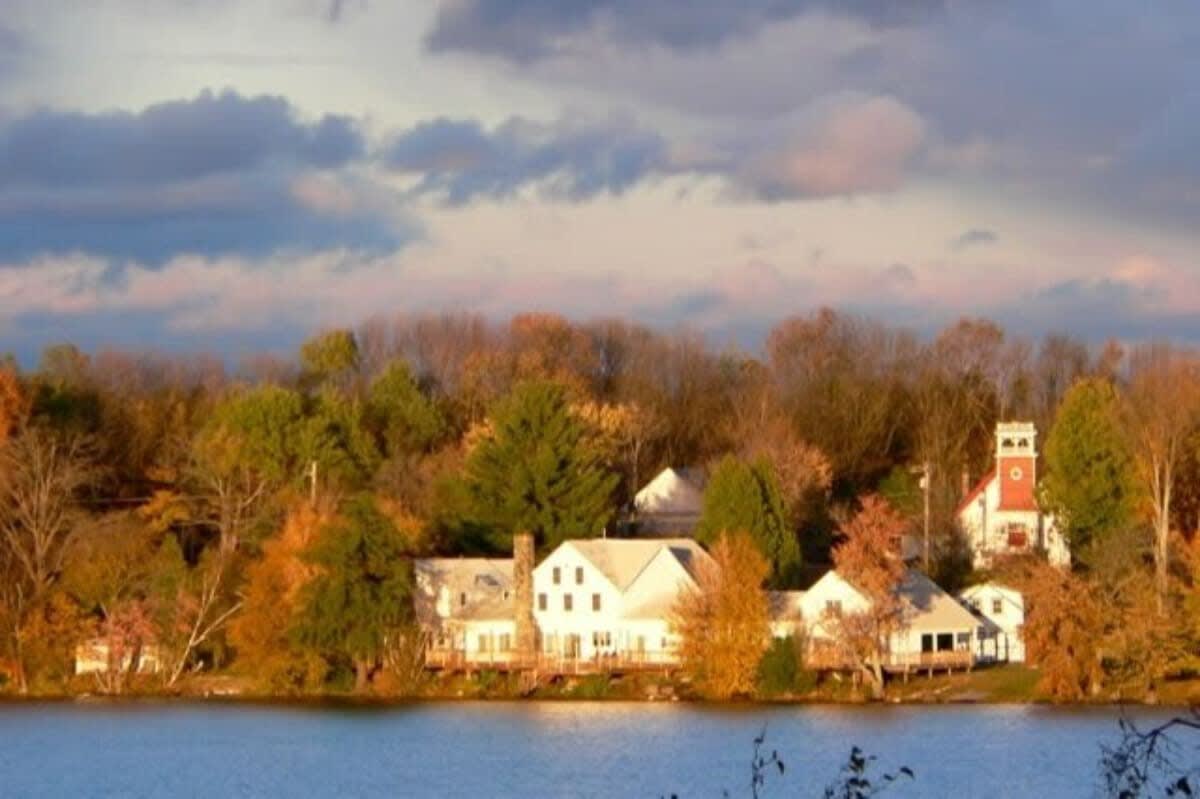 Panoramic view of the lakefront property and surrounding area during autumn, showing the white house nestled among colorful trees with a church steeple visible in the distance.