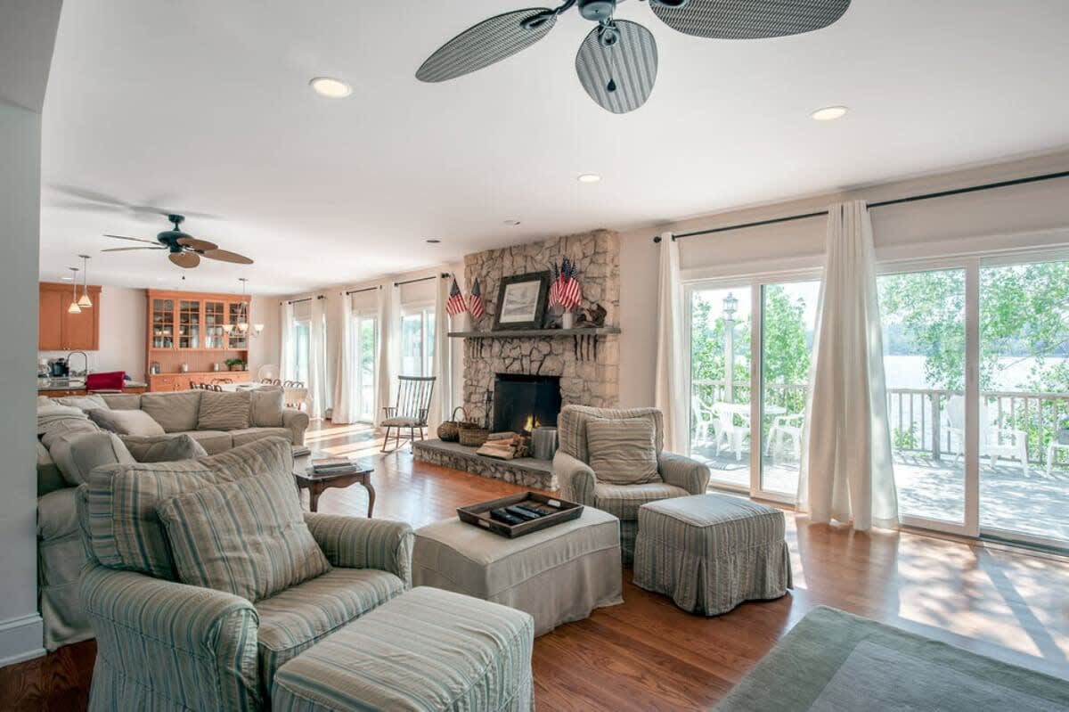 Bright and spacious living room featuring a large stone fireplace, comfortable striped armchairs, and a light-colored sectional sofa, with large sliding glass doors opening to a deck and lake views.