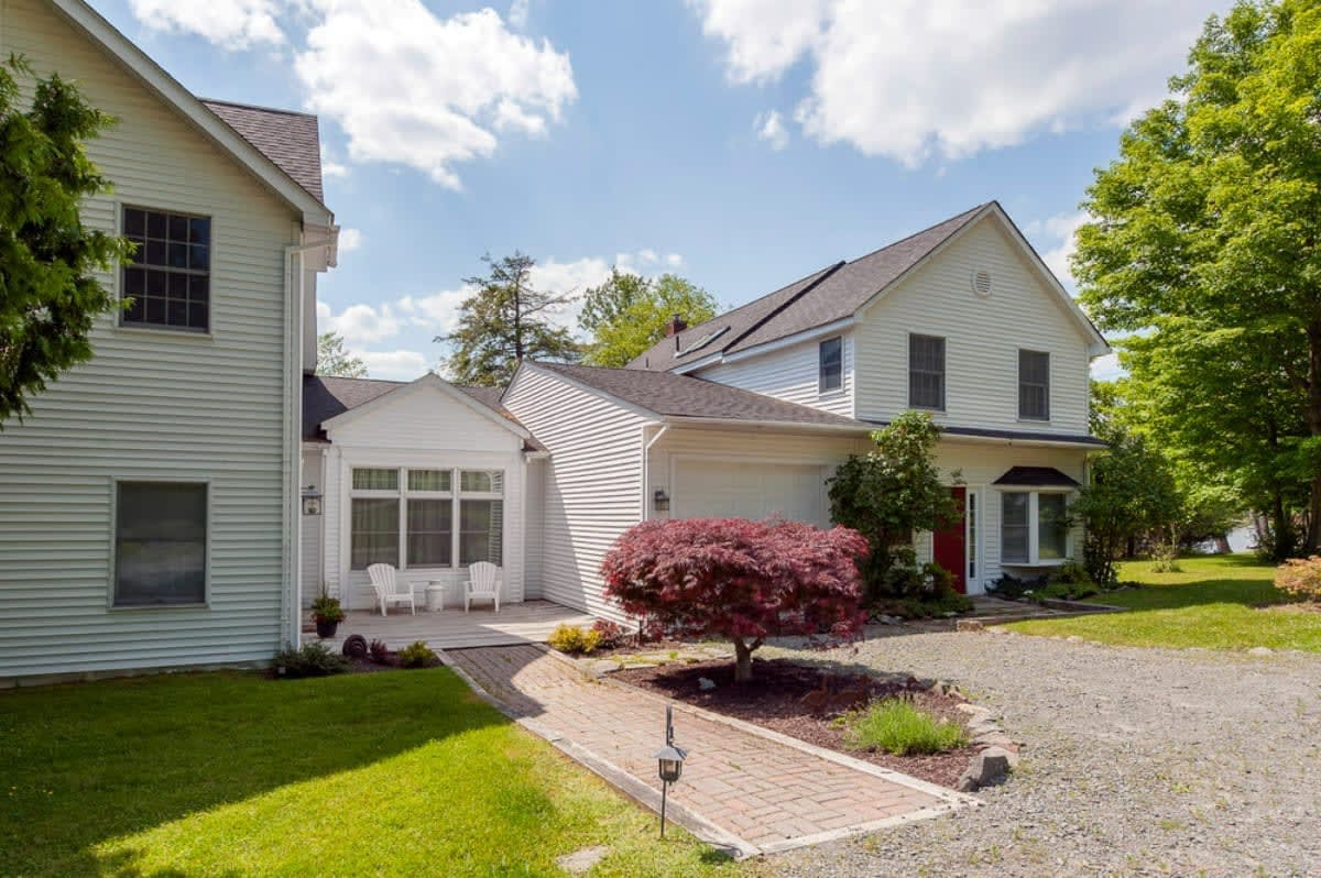 A view of the white-sided house from the front, showing a paved brick walkway leading to the entrance, a gravel driveway, and a vibrant red Japanese maple tree in the foreground.