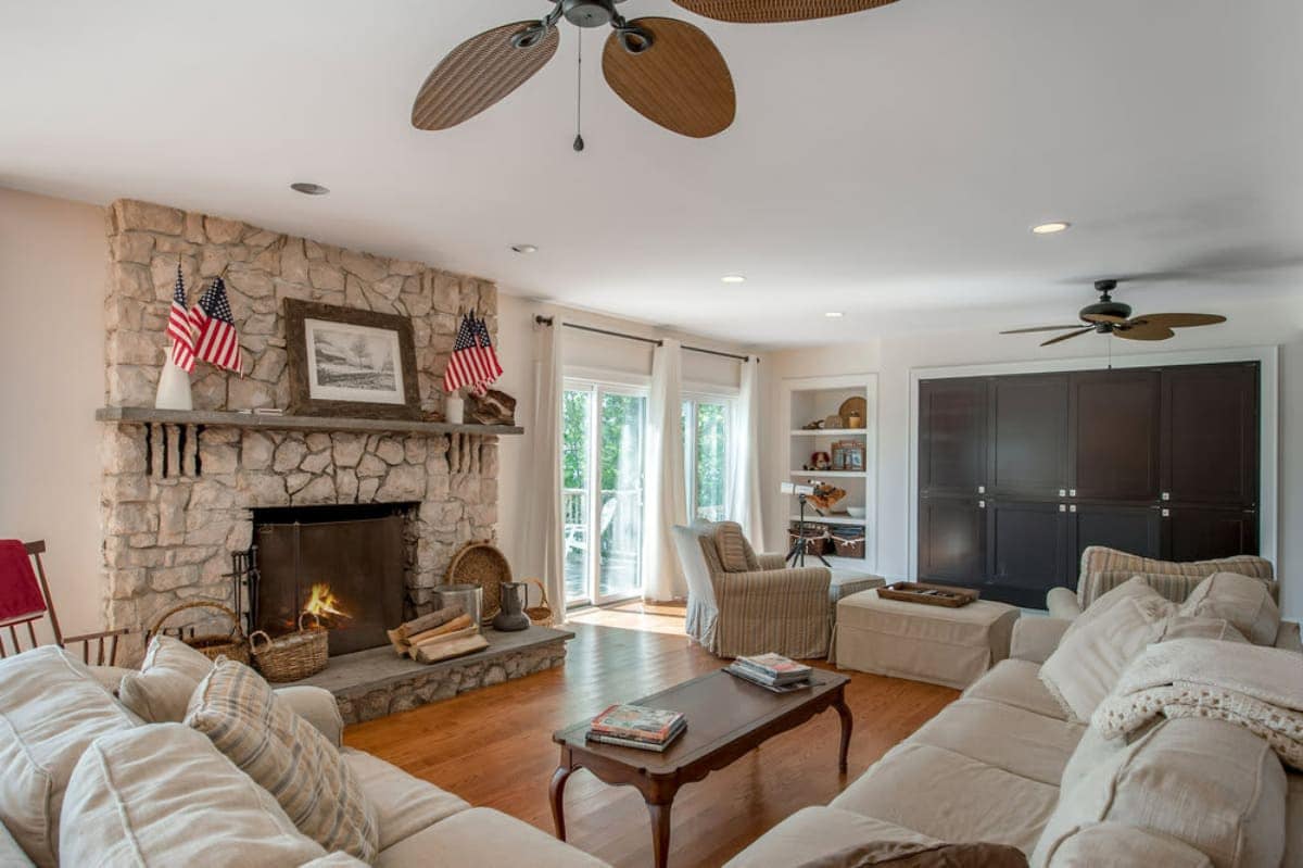 Cozy living room featuring a large stone fireplace with a mantel adorned with American flags, light-colored sofas, and warm hardwood floors. Two ceiling fans provide air circulation.