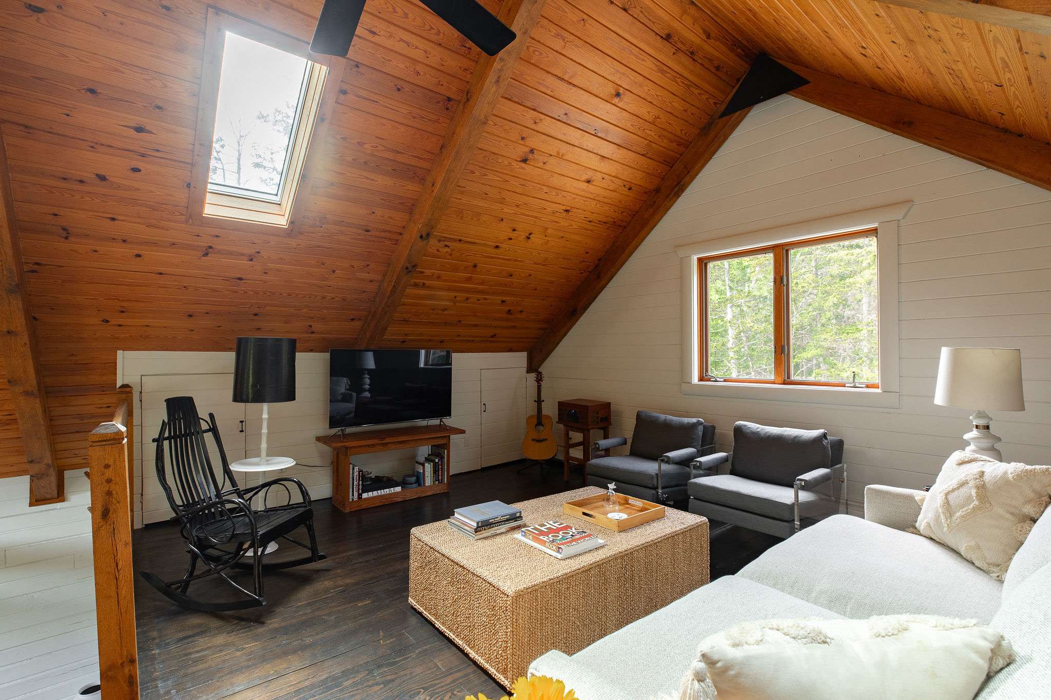 Cozy upstairs living area with a vaulted wooden plank ceiling and a skylight, featuring a TV console, a black rocking chair, and a light-colored sectional sofa around a woven coffee table. A window offers views of trees.