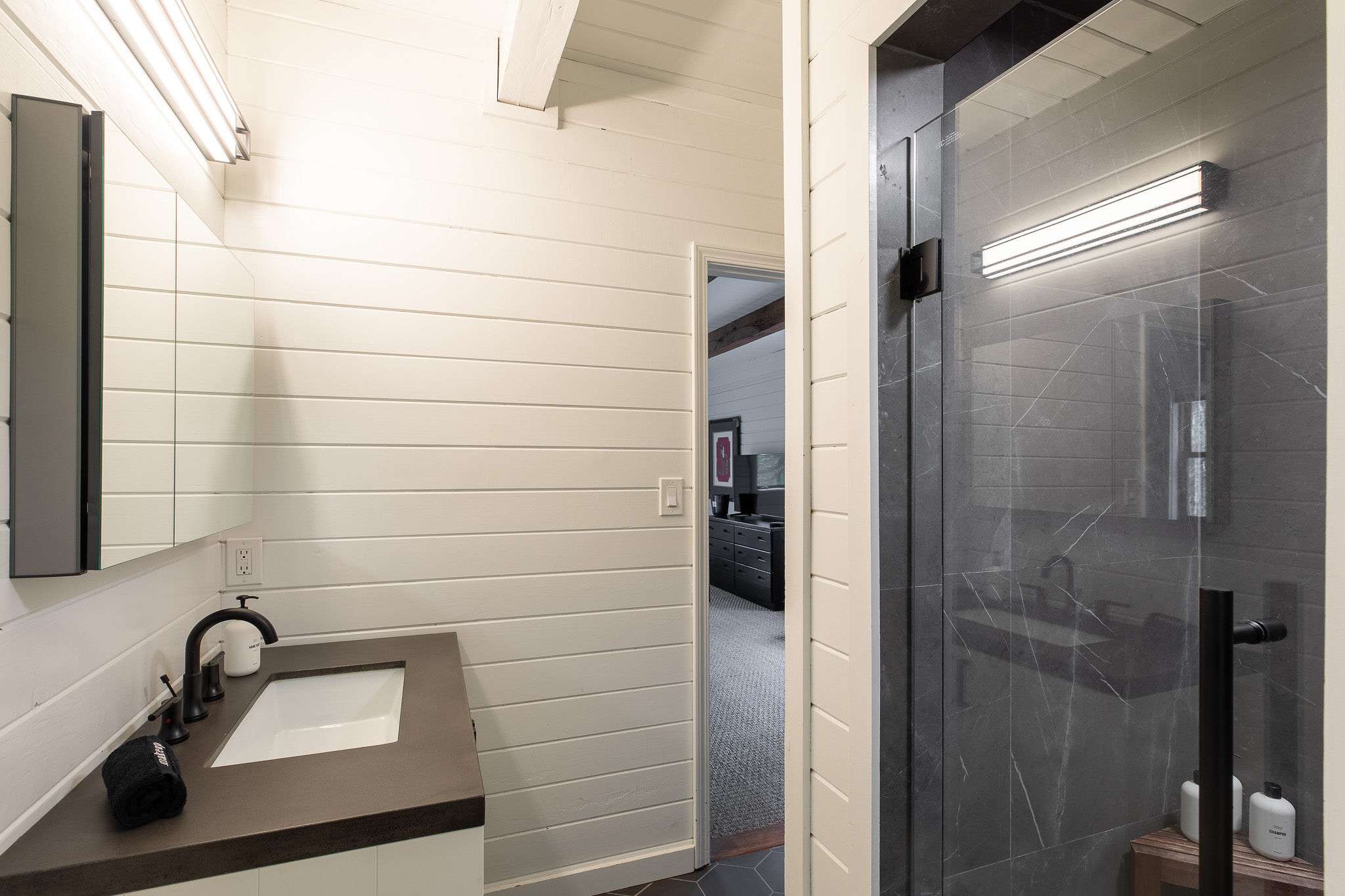 View of the modern bathroom showing the vanity with white shiplap walls and a dark countertop, adjacent to a sleek shower with dark grey marble-patterned tiles and a glass enclosure. A doorway leads to a bedroom.