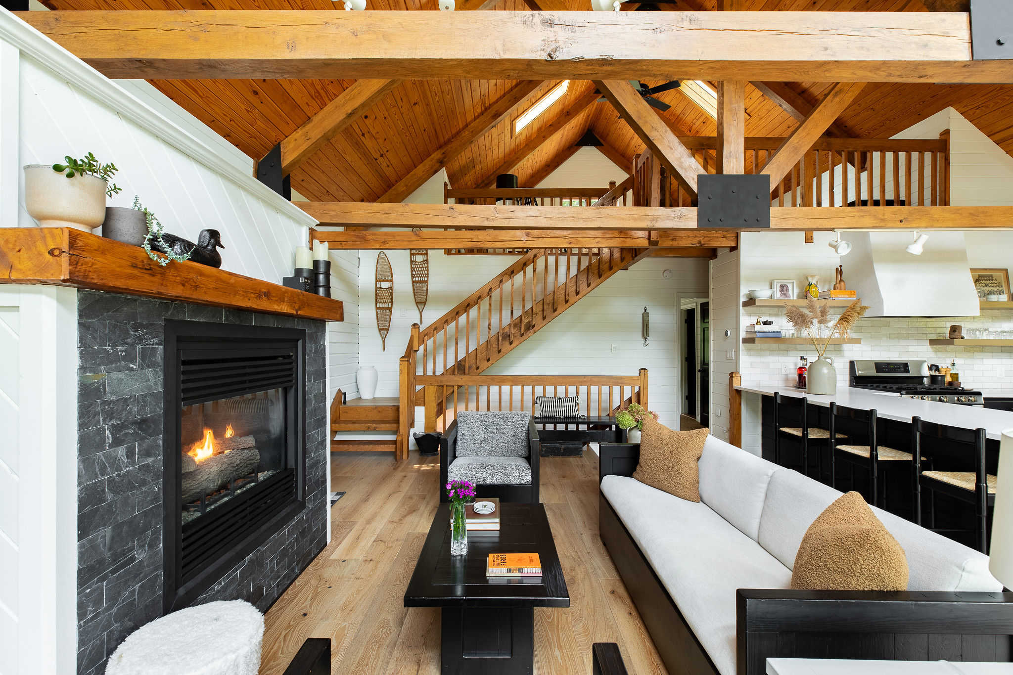 Spacious open-concept living area with a modern fireplace, white sofa, and exposed wooden beams, leading to a kitchen and a wooden staircase.