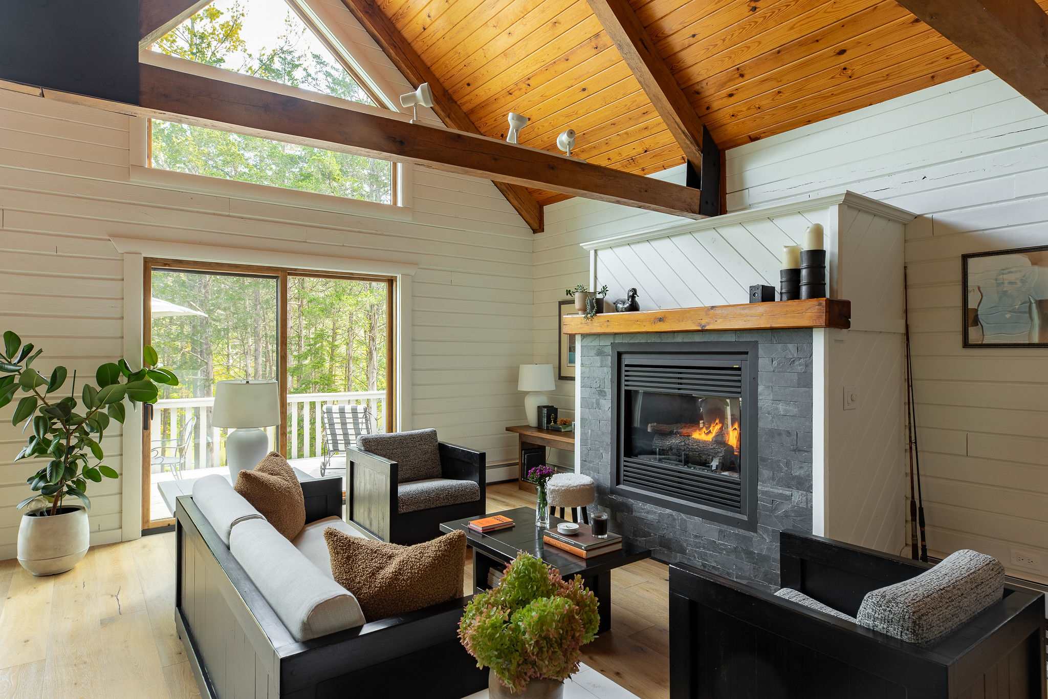 A bright living room features a modern fireplace with a dark stone surround and a rustic wooden mantel, set against white shiplap walls. Large windows and a sliding glass door offer views of the surrounding trees, complementing the light wood floors and vaulted ceiling with exposed beams.