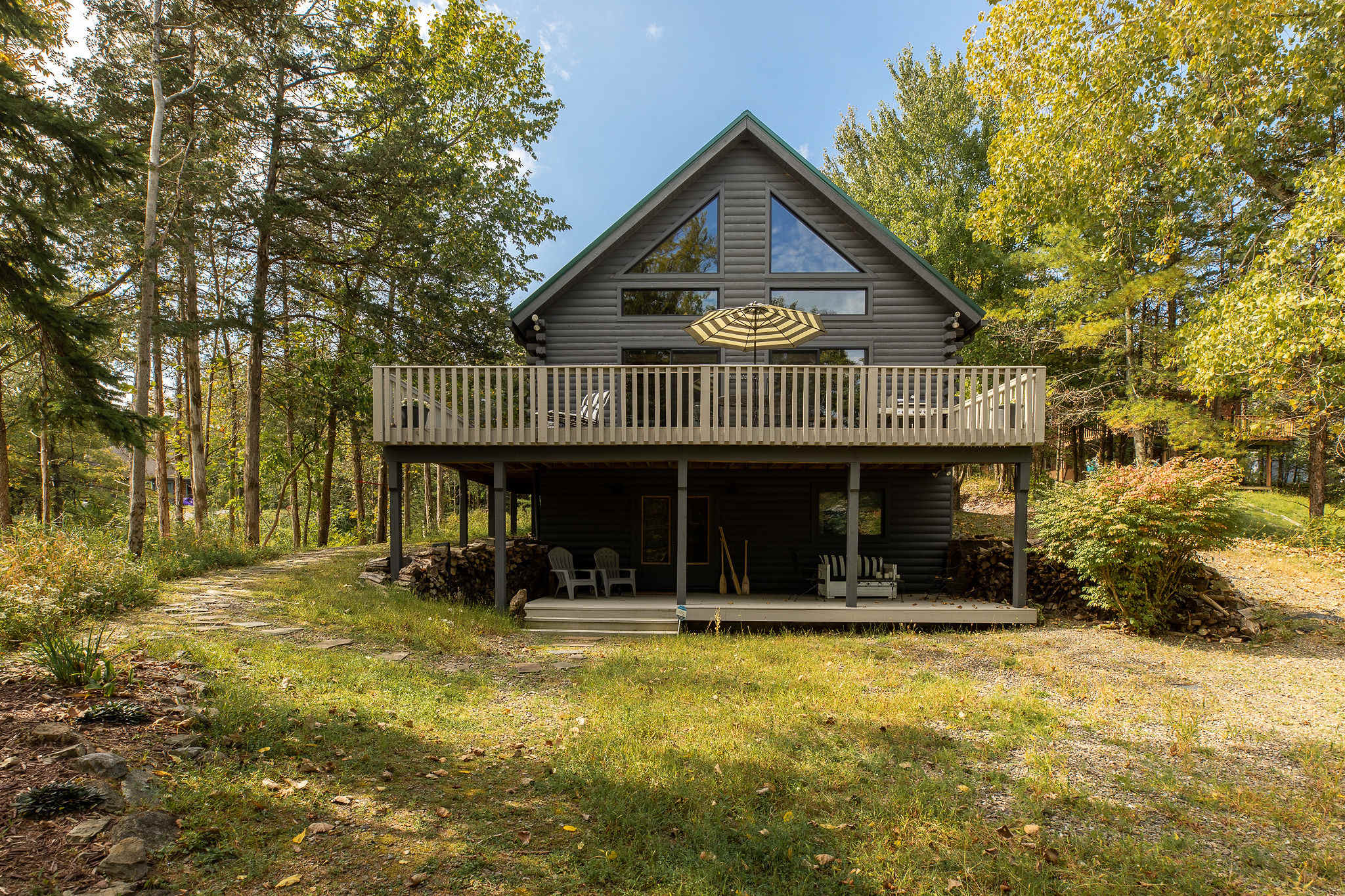 The back of the gray log cabin features a large elevated wooden deck with outdoor seating and a striped umbrella, and a lower-level entrance with stacked firewood underneath, surrounded by trees.