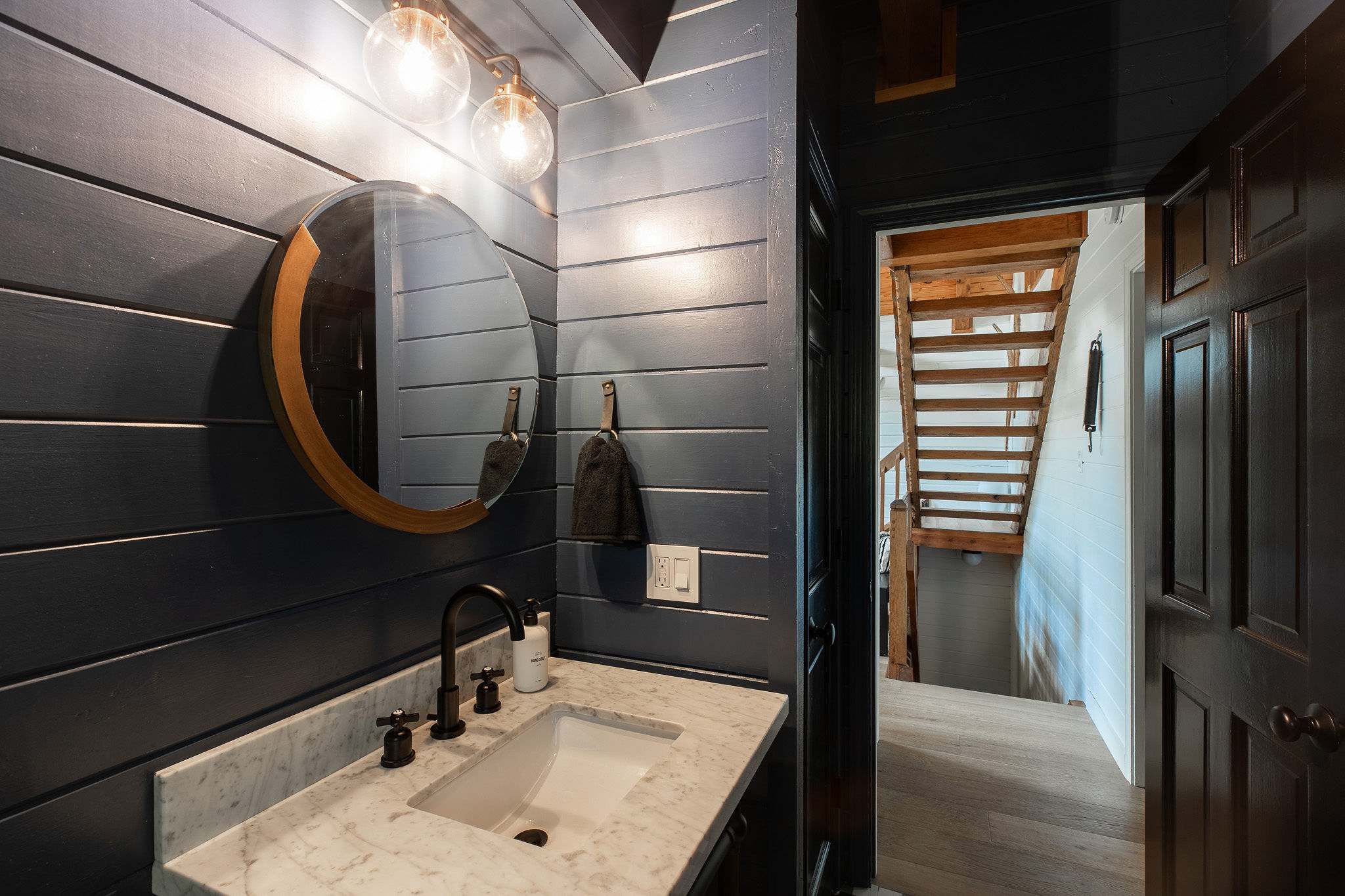 Stylish half-bathroom with dark blue shiplap walls, a white marble vanity, and a round mirror, featuring a black faucet and industrial-style light fixtures.