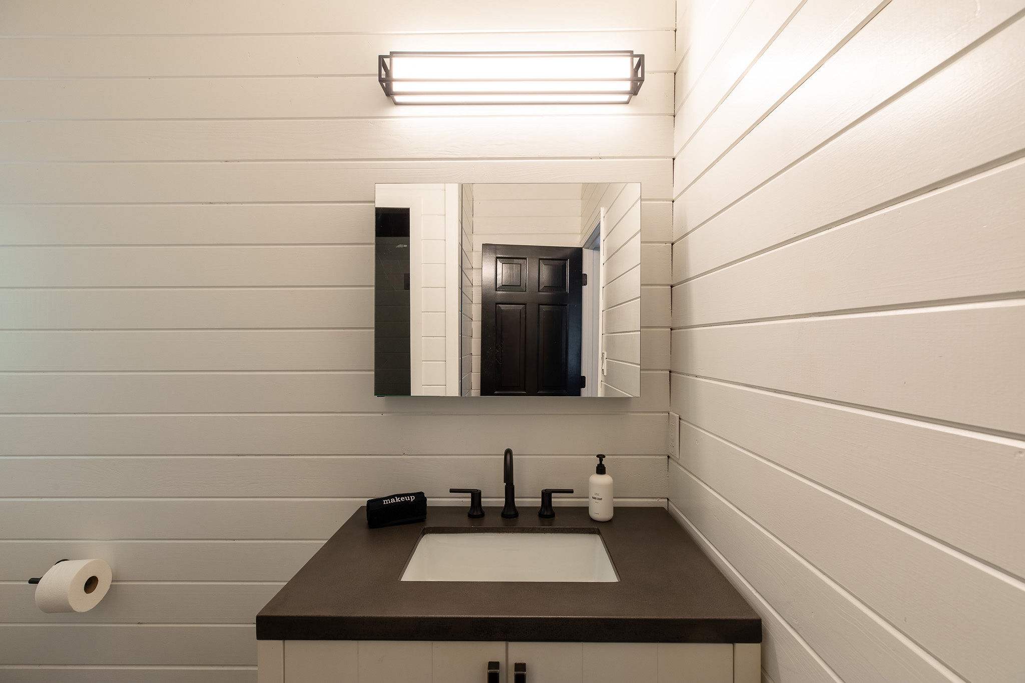 Modern bathroom vanity with white shiplap walls, a dark countertop, and a rectangular white sink with a black faucet. A large mirror is illuminated by a linear light fixture above.