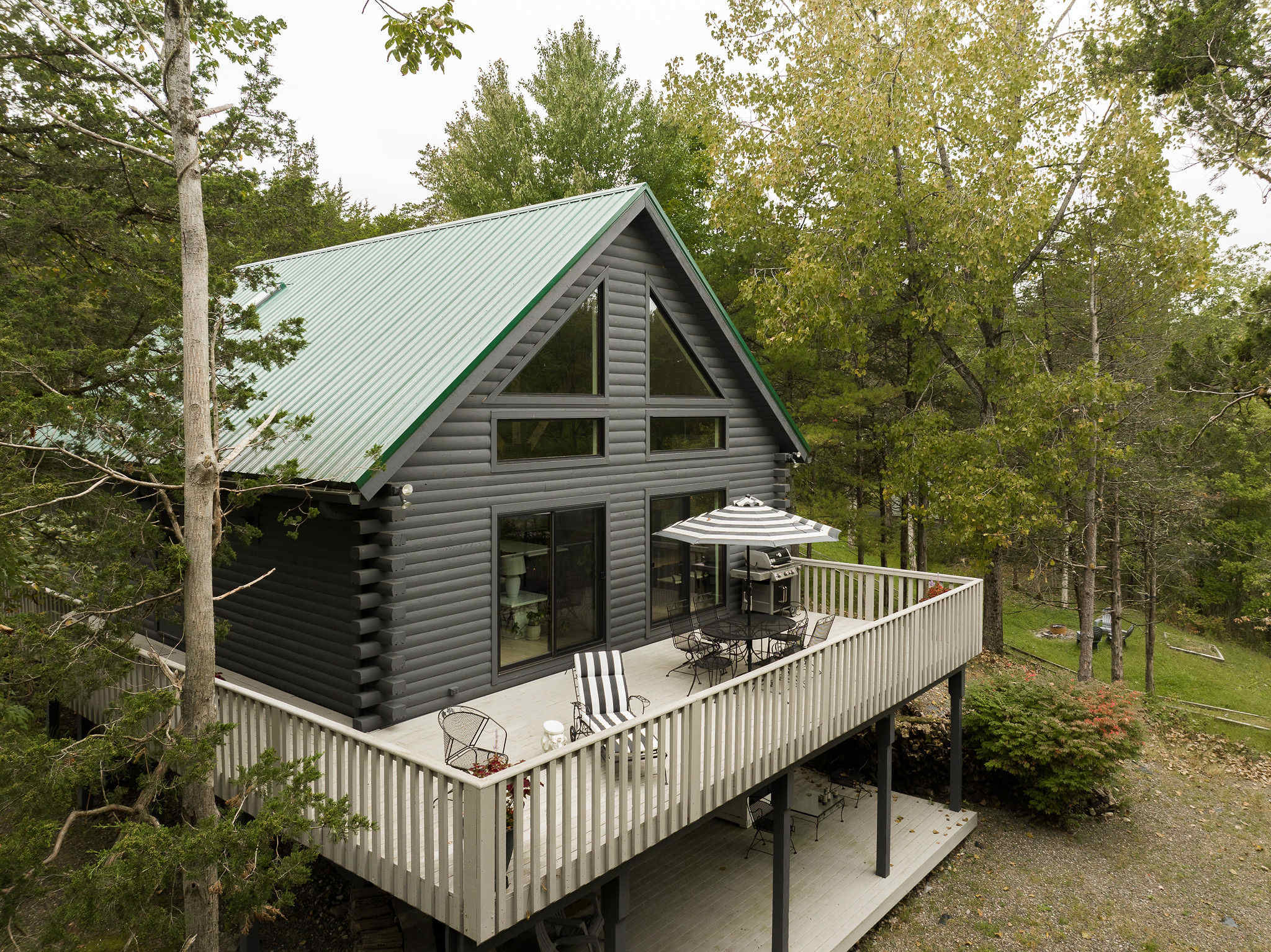Aerial view of the back of the gray log cabin, highlighting a spacious wooden deck furnished with outdoor seating and a BBQ grill, offering an elevated view of the surrounding wooded area.
