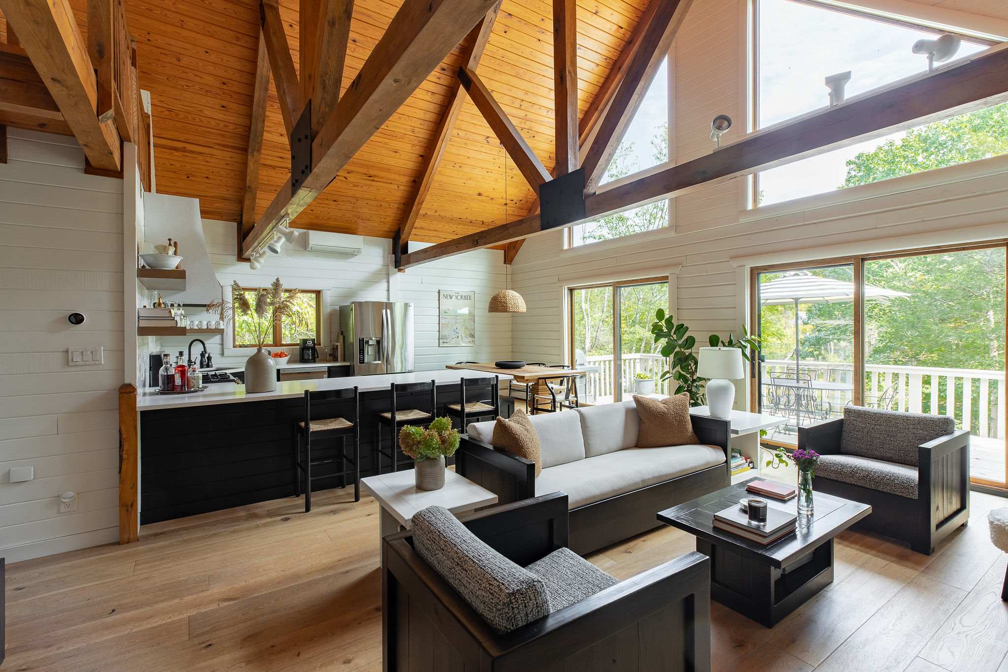 A spacious open-plan living room and kitchen area features a large white kitchen island with bar seating, facing a comfortable sofa and armchairs. The room is characterized by its vaulted wooden ceiling with exposed beams, white shiplap walls, and light hardwood floors, with large windows offering outdoor views.