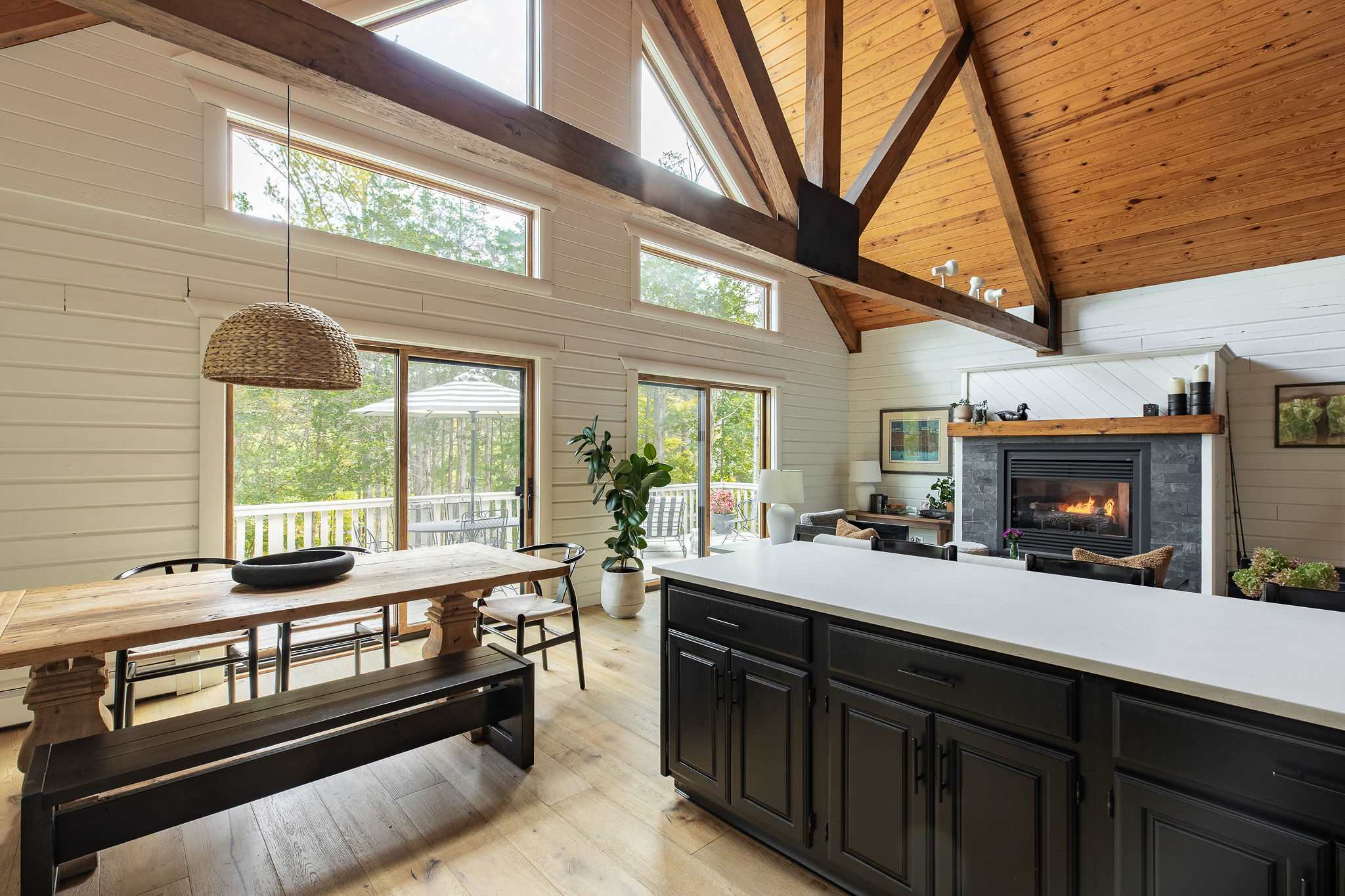 Bright open-concept living and dining area with vaulted wooden ceilings, exposed beams, and large windows overlooking a wooded deck, featuring a fireplace and a kitchen island.