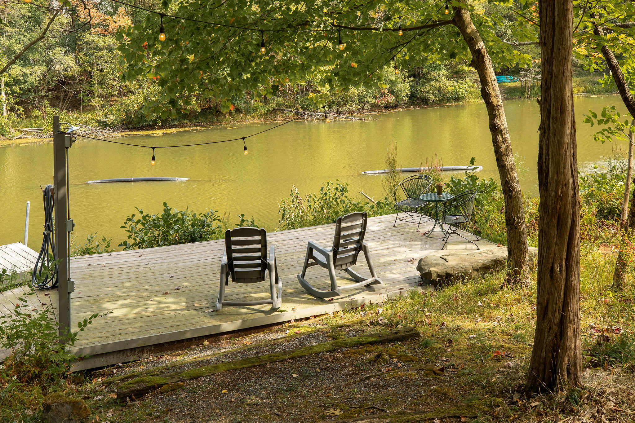 A wooden dock extends over the calm lake, featuring two modern rocking chairs and a small bistro set, illuminated by string lights overhead, offering a serene spot by the water.
