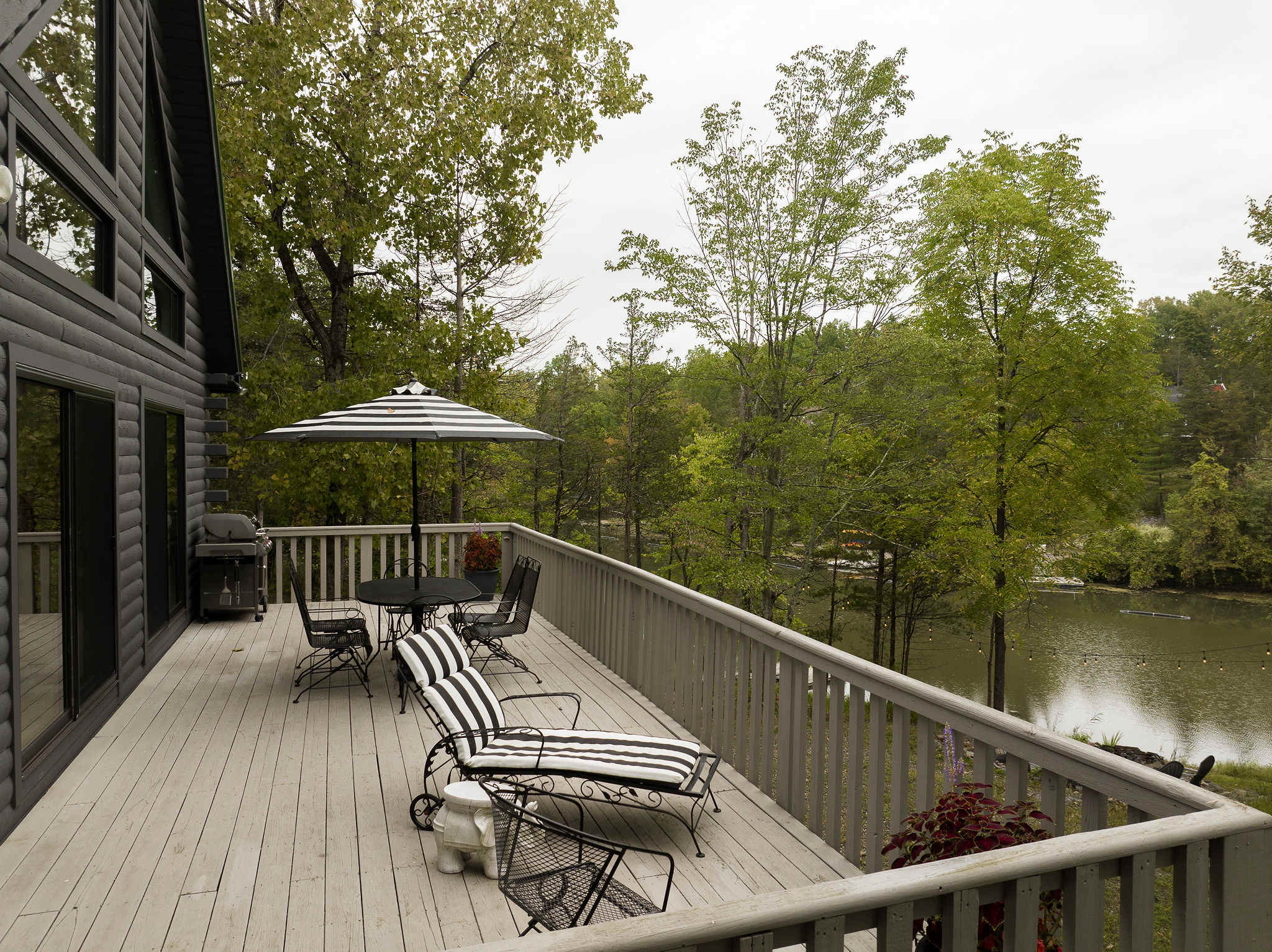 A spacious wooden deck extends from the cabin, furnished with a black metal dining set under a striped umbrella and a chaise lounge. The deck offers a scenic view of the surrounding trees and a glimpse of the lake in the distance.