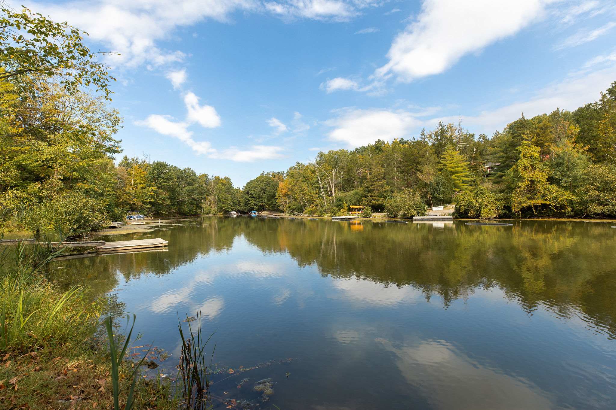 A wide shot of a serene lake reflecting a blue sky with fluffy white clouds, surrounded by lush green trees. Several small docks and boats are visible along the tree-lined shore, highlighting the tranquil waterfront setting.