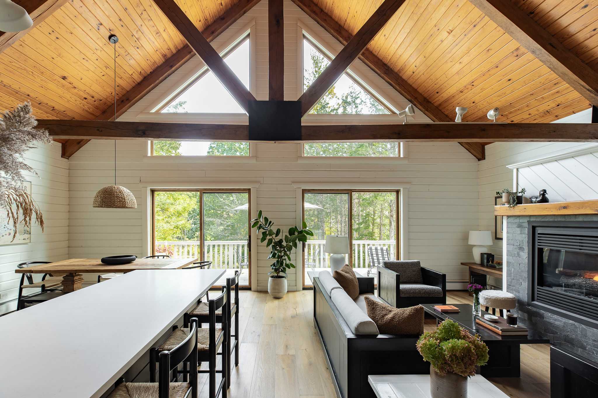 An open-concept living and dining area boasts a high vaulted ceiling with exposed wooden beams and a light wood finish, complemented by white shiplap walls. Large A-frame windows and sliding glass doors provide abundant natural light and views of the surrounding nature.