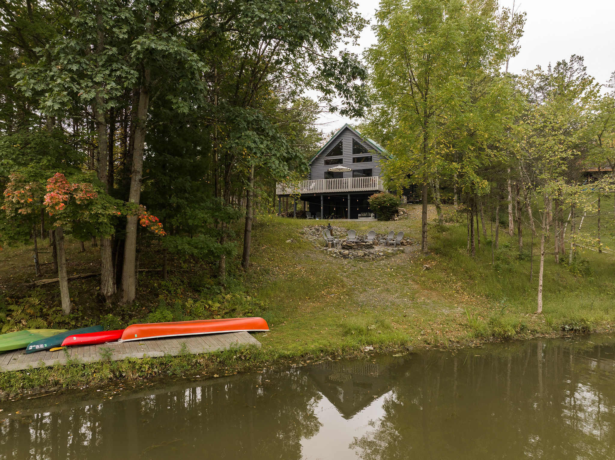 Expansive aerial view of the property, showing the gray log cabin, the fire pit area, and the lake with canoes pulled onto the shore, all nestled among trees, emphasizing its secluded lakefront appeal.