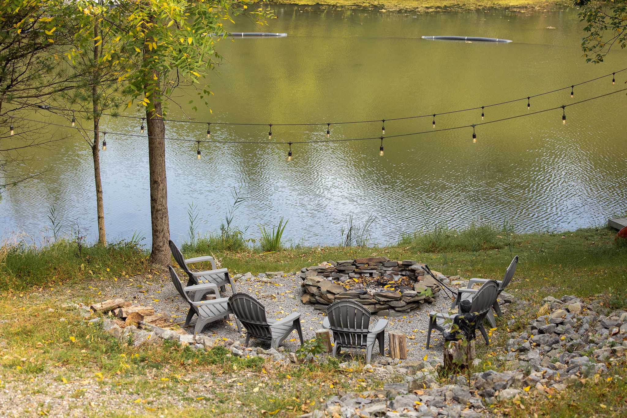 A cozy stone fire pit surrounded by several gray Adirondack chairs sits on a gravel patch near the lake, with string lights strung above, creating an inviting evening ambiance.