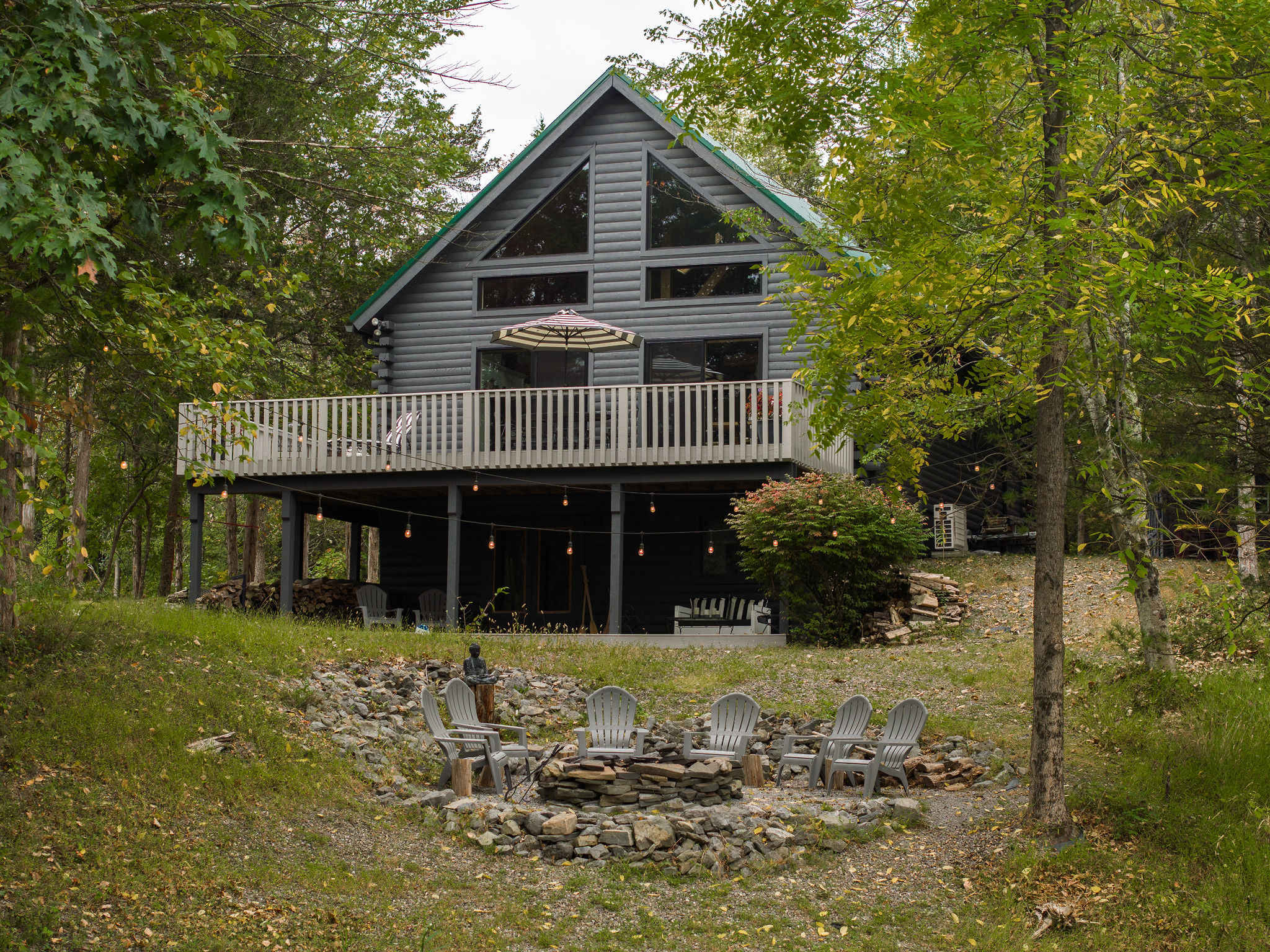 The gray log cabin stands amidst green trees, featuring a large elevated wooden deck with outdoor furniture and an umbrella. Below the deck, a stone fire pit with Adirondack chairs is set on a grassy slope, offering a view towards the lake.