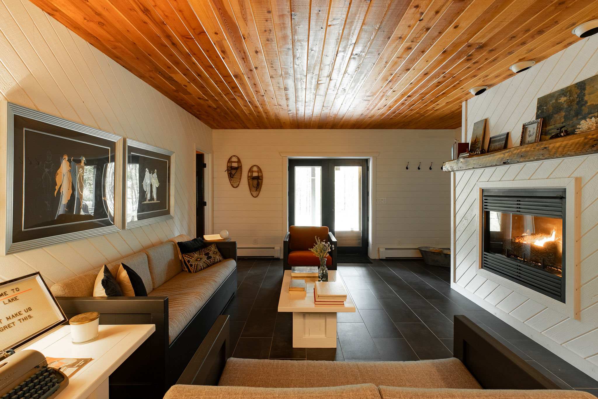 Inviting living room with a long sofa, dark tile floors, and a modern fireplace, featuring white shiplap walls, a warm wooden ceiling, and framed artwork.