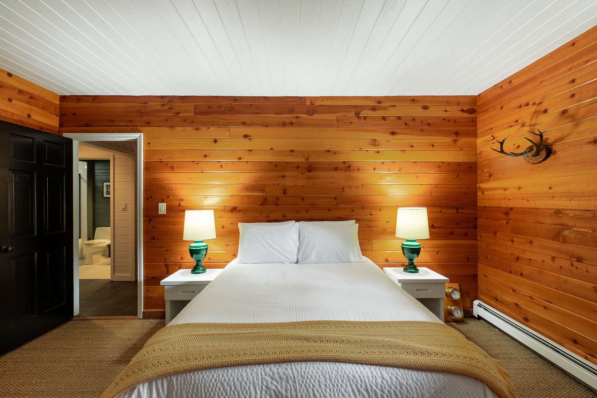 Warm bedroom featuring horizontal wood-paneled walls and a white plank ceiling. A queen-sized bed with white linens and a yellow throw is flanked by white nightstands with green-based lamps. A doorway leads to a bathroom.
