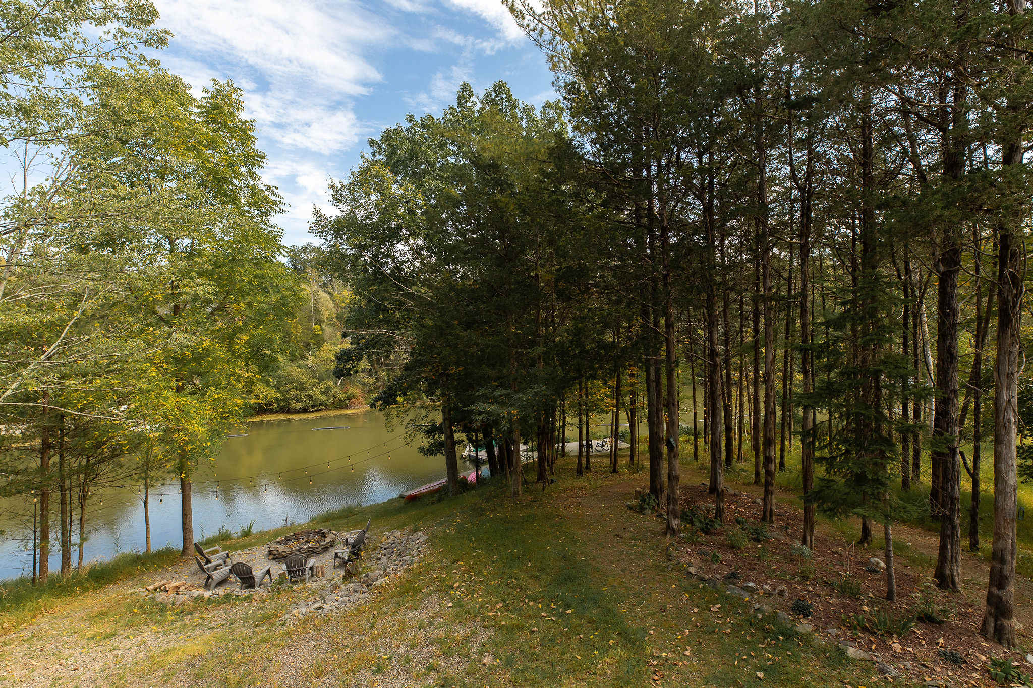 Aerial view showcasing the property's lakefront setting with a fire pit area, outdoor seating, and dense surrounding trees under a partly cloudy sky, highlighting the natural landscape.
