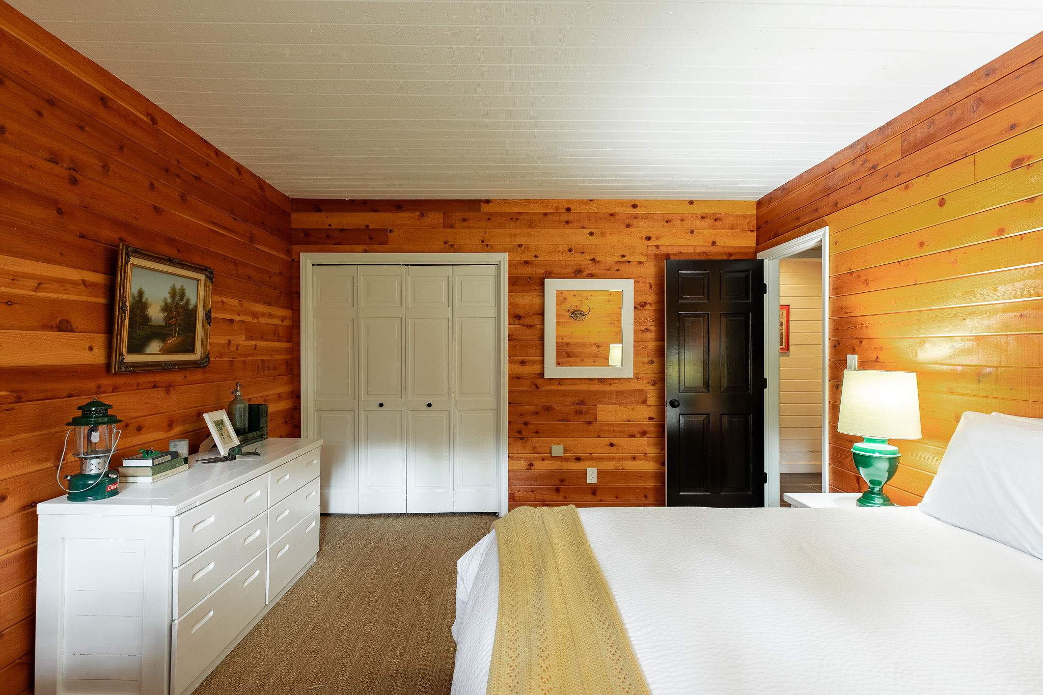 Another view of the wood-paneled bedroom, showcasing a white dresser with decor, a white bi-fold closet, and a black interior door. The room maintains its warm, rustic aesthetic with a white plank ceiling.